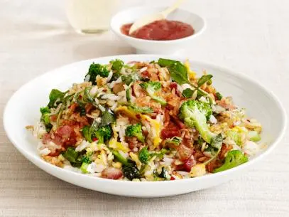 A fried rice, vegetables and bacon mixture in a white and gray bowl on a tan table setting
