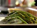 A close up of asparagus that has been baked in an oven. The asparagus is sitting on a white and black plate. There is steam coming off of the asparagus. The asparagus is bright green.