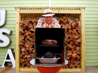 Alton Brown stands behind his homemade smoker that he is using to cook his pork shoulder.