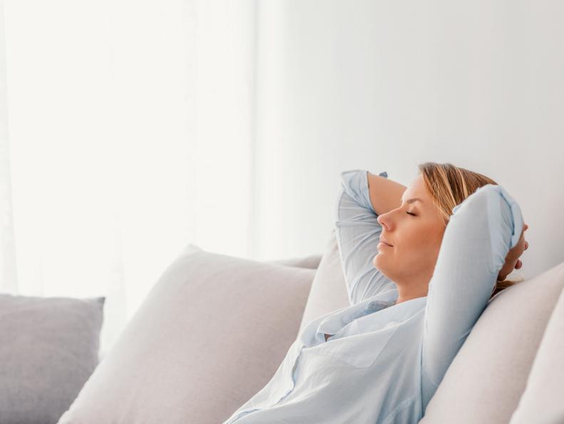 Side view portrait of a relaxed happy woman, with eyes closed,  resting sitting on a couch  at home during bright day. Portrait of a Caucasian young, brown hair girl relaxing on a sofa after work at home sitting on a sofa in the living room at home with a warm light of sunset, seeping with hands behind head.
