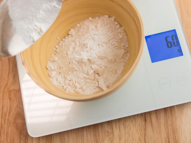 Kitchen scale with tarred bamboo bowl filled with 60 gram of flour. Out of focus spoon in foreground. Wooden board. Light effect. High point of view.