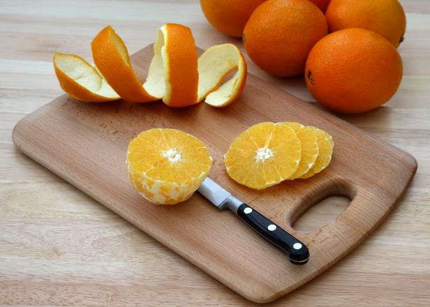 96621711 Peeled orange being sliced on chopping board beside coil of orange peel removed from orange. Chopping board on kitchen table with a few more oranges in the background.
