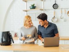 Beautiful young caucasian wife in kitchen cooking healthy oil free meal in modern air fryer with handsome husband working on laptop in kitchen at home