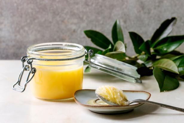 Homemade Melted ghee clarified butter in open glass jar and spoon on saucer standing on white marble table with green branch. Grey wall at background. (Photo by: Natasha Breen/REDA&CO/Universal Images Group via Getty Images)
