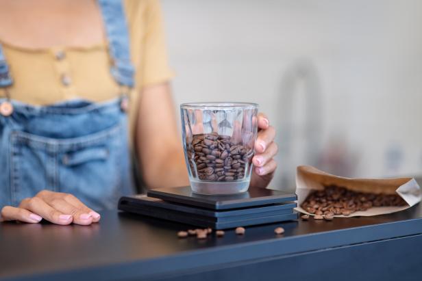 Whole bean coffee. Womans hand putting a glass with coffee beans on scales on counter, no face is visible