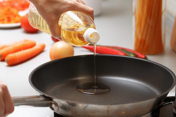 Home cooking. A woman pouring oil from a bottle into the pan in the kitchen, near fresh vegetables and pasta.