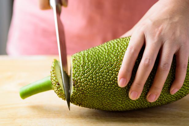 Hand holding kitchen knife and cutting young jackfruit on wooden board for cooking