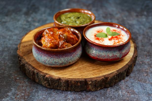 Stock photo showing close-up view of bowls containing raita (yoghurt based sauce), mint coriander chutney and green mango pickle on a wooden chopping board against a grey background.