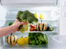 cropped shot of female hand hold fresh broccoli near open refrigerator full with different vegetables and fruits inside, healthy eating concept