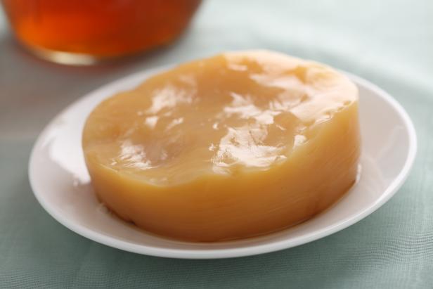 A kombucha scoby (symbiotic colony of bacteria and yeast) resting on a plate, with a jar of kombucha tea in the background