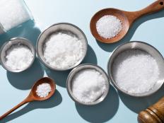 A Variety of Sea Salt Including Fine Salt, Kosher Salt, Coarse Salt, Flake Salt in Bowls and Spoons on Light Blue Background Directly Above View.