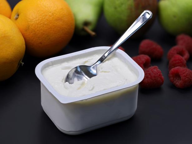 quark or cream cheese in plastic box with spoon on black backround with fruits, healthy nutrition for breakfast.