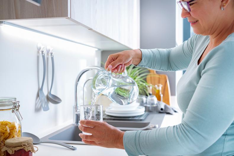 Side view of smiling mature woman pouring drinking water from jug to a glass in the kitchen. High resolution 42Mp indoors digital capture taken with SONY A7rII and Zeiss Batis 40mm F2.0 CF lens