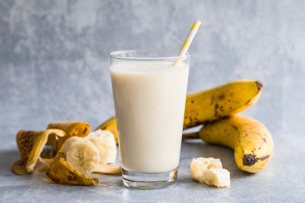 Banana smoothie of organic bananas. There is a high glass filled with banana smoothie in the centre, and bananas and banana peels around the drink. With an environmentally friendly paper straw.
