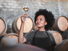Black woman tasting wine at a winery, looking and checking the color and quality of the years produce. Young African American sommelier proud of the new addition, analyzing white wine in a cellar