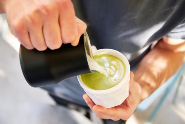 Male barista making vegan matcha cappuccino with oat milk with latte art in outdoor cafe.