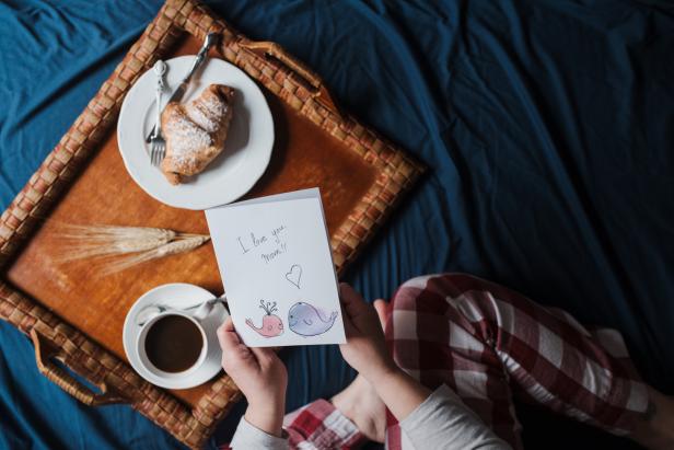 View from above of a mother holding a handmade Mother's Day card with a tasty breakfast in bed. She is on a blue colored sheet.