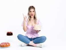 Host Meghan Rienks sitting on the floor, she has a surprise look on her face, holding her confetti cake in the left hand, while her chocolate cake, and bun cake is on the left,  wearing her pink top and jeans, as seen on Food Network’s Just Ask The Baker, Season 1 (Horizontal)