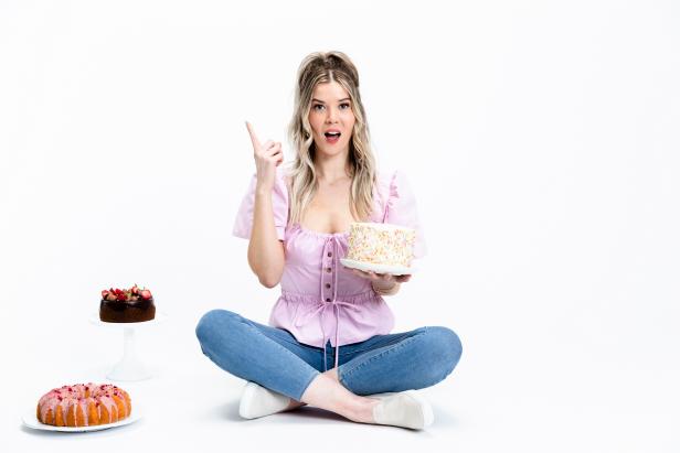Host Meghan Rienks sitting on the floor, she has a surprise look on her face, holding her confetti cake in the left hand, while her chocolate cake, and bun cake is on the left,  wearing her pink top and jeans, as seen on Food Network’s Just Ask The Baker, Season 1 (Horizontal)