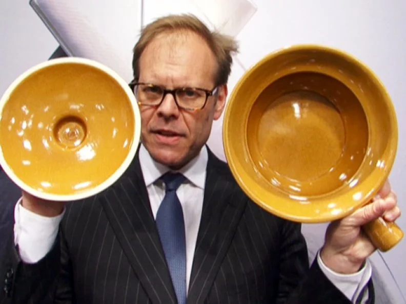 Alton Brown wearing a pinstriped blue suit while holding a ceramic pot and lid