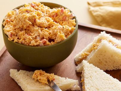Three stacked crustless Pimiento Cheese sandwiches on a plate beside a bowl full of Pimento Cheese