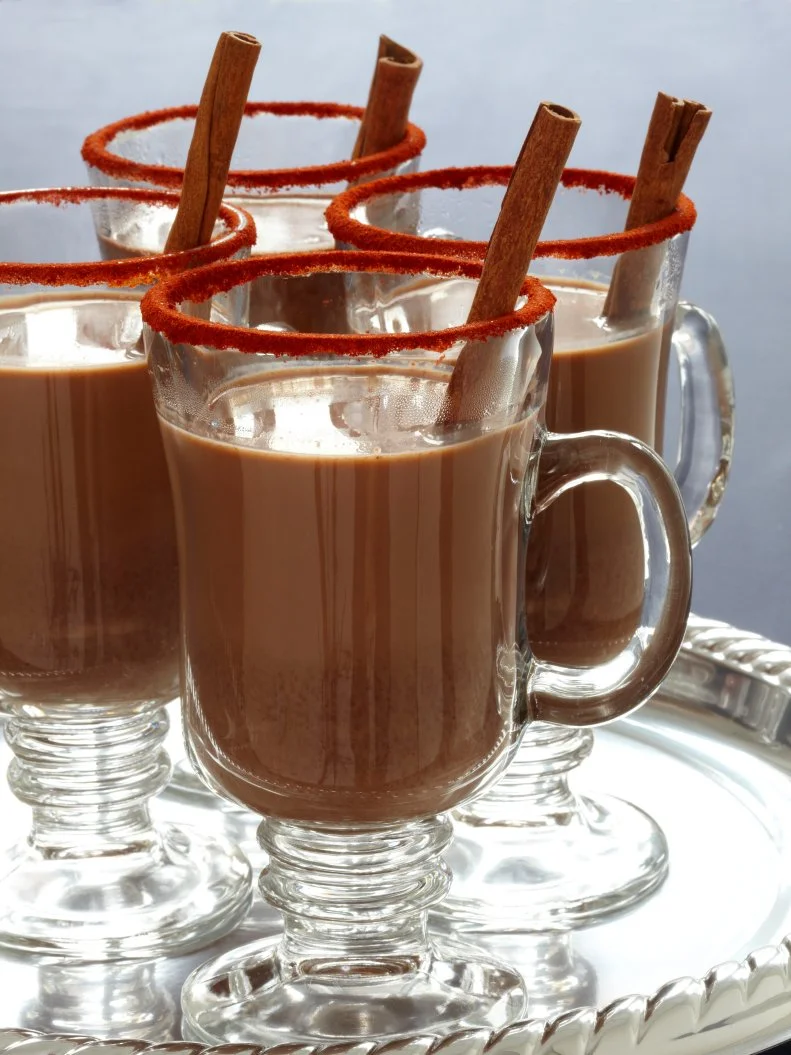 Four glass mugs of Mexicocoa garnished with cinnamon sticks and sugar around the rims