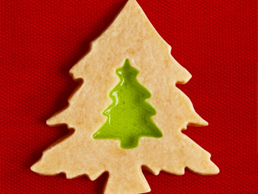 A christmas tree shaped cookie with a translucent green window pane candy in the middle and placed against a red fabric background 