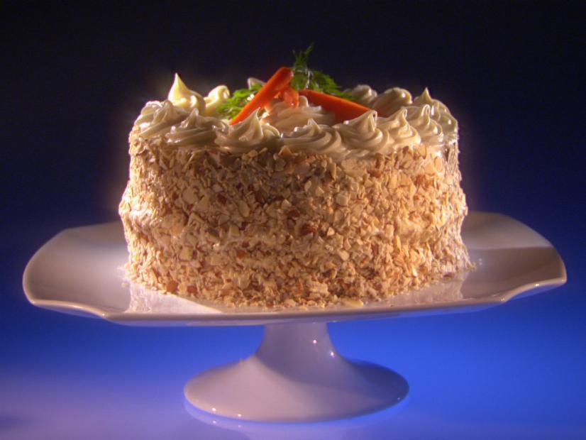 A Cardamom Carrot Cake topped with frosting carrots atop a white dessert pedestal