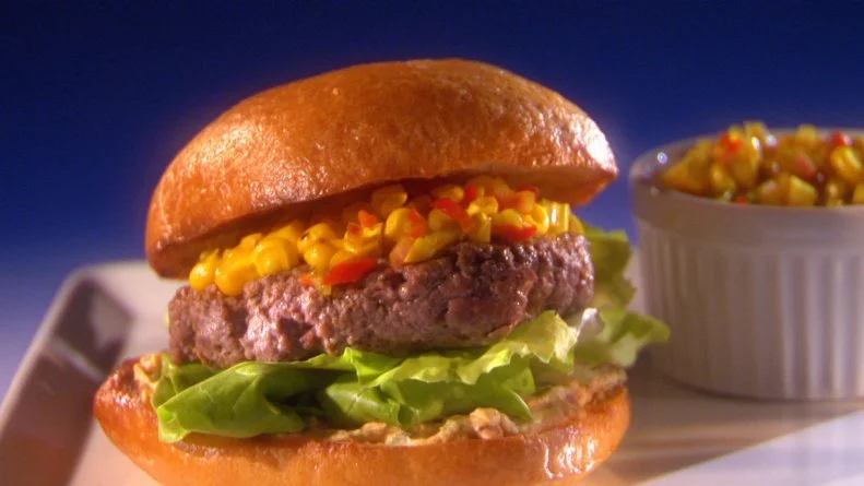 A hamburger on a bed of lettuce and topped with corn next to a container of corn 