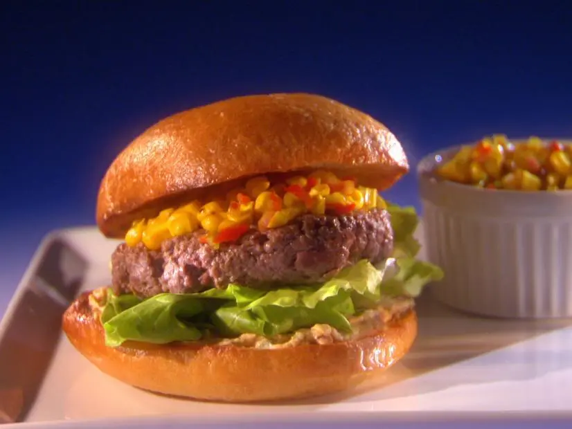 A hamburger on a bed of lettuce and topped with corn next to a container of corn 