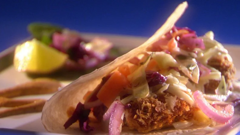 A catfish taco on a thin square gray plate with a lemon wedge and some drizzled sauce