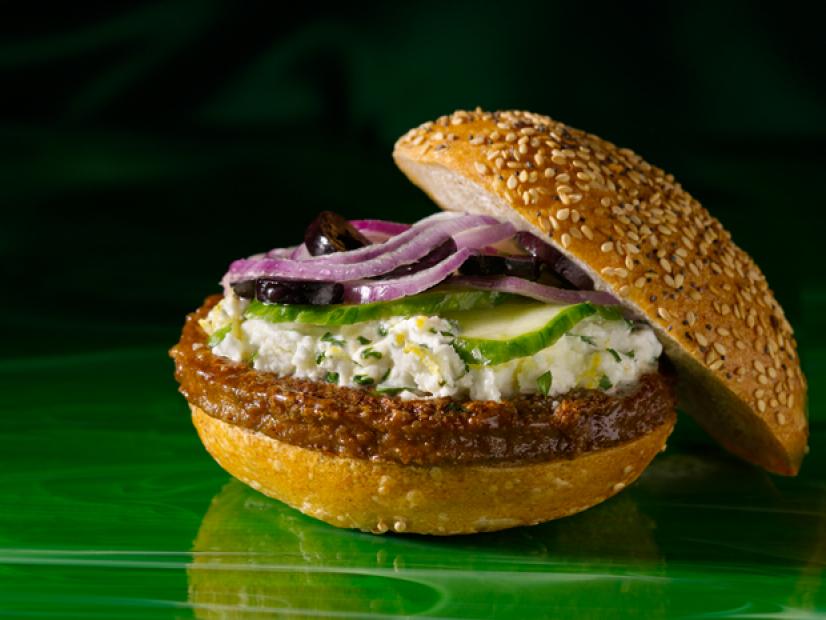 A burger on a sesame seed bun with a Greek sauce, black olives and red onions