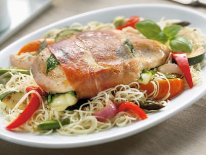 A chicken breast wrapped with pork on a bed of noodles with sliced vegetables