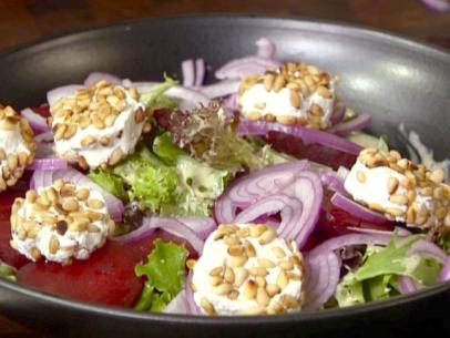 A beet salad with goat cheese topped with pine nuts is served with slices of red onion, baby greens, and baby spinach.