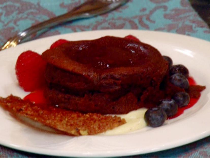 A chocolate molten love cake is served on a bed of creme anglaise, topped with chocolate sauce, and garnished with blueberries, raspberries, pistachio tuile, and raspberry coulis.