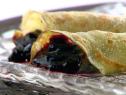 Two blueberry jam filled crepes sprinkled with confectioners sugar on a plate against a white background