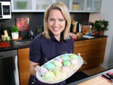 Melissa darabian standing a kitchen and wearing a dark purple dress while hold a platter full of pastel colored Easter eggs