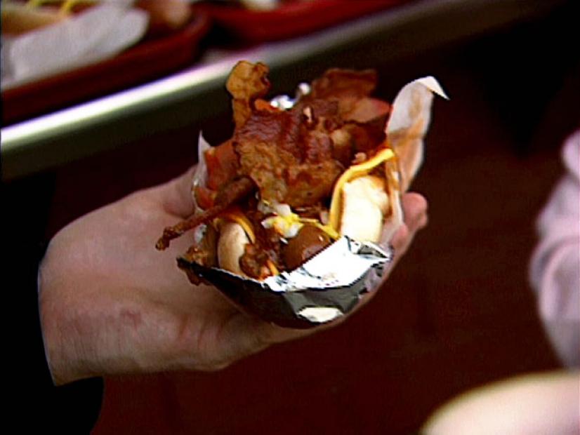 Bobby Flay holding a hot dog with cheese and topped with bacon