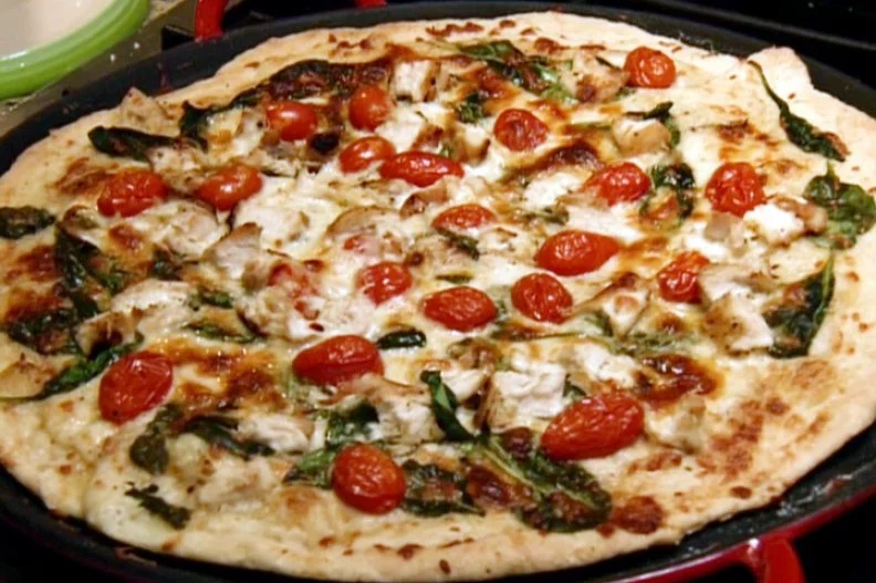 Chicken alfredo pizza is topped with red and yellow grape tomatoes, baby spinach, grated mozzarella, and parmesan.
