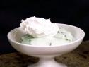 A scoop of mint chocolate chip ice cream in a white bowl is topped with whipped cream.