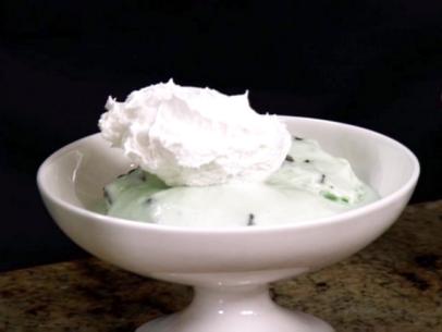 A scoop of mint chocolate chip ice cream in a white bowl is topped with whipped cream.