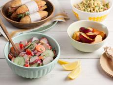 Potato Salad in a light green dish on a white washed tabletop with dishes of slice fruite, sandwiches and macaroni salad