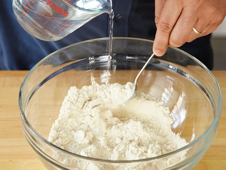 Water being poured from a measuring cup into a mixing bowl full of flour