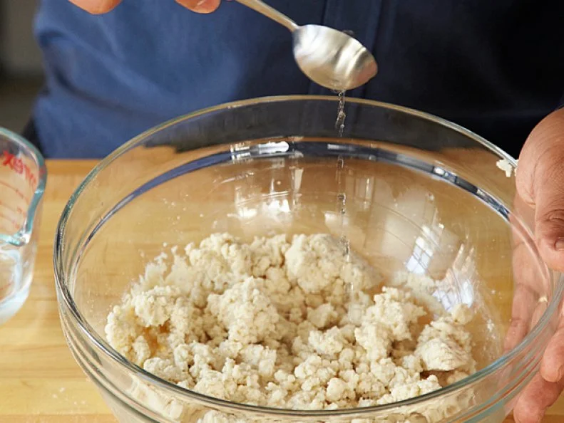 Water being poured from a measuring spoon into a clear glass bowl containing dough