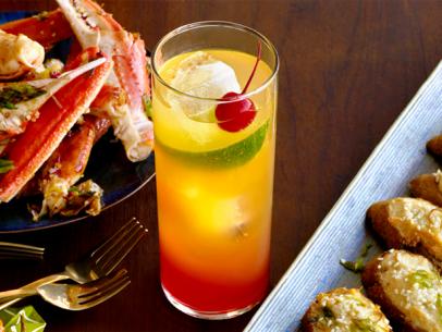 A red and yellow cocktail with a cherry, lime and lemon wedges in a tall slinder glass in between dishes of crabs legs and bruschetta
