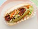A grilled hot dog topped with lettuce, carrots and a pickle wedge on a bun placed in a paper hot dog wrap