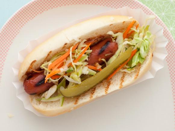 A grilled hot dog topped with lettuce, carrots and a pickle wedge on a bun placed in a paper hot dog wrap