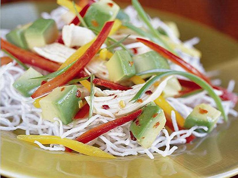 A salad made of chopped avocados, noodles, and red and yellow peppers place in a golden bowl placed on a wooden surface