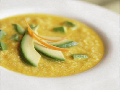 A yellow soup containing chunks and slices of avocado in a simple white bowl