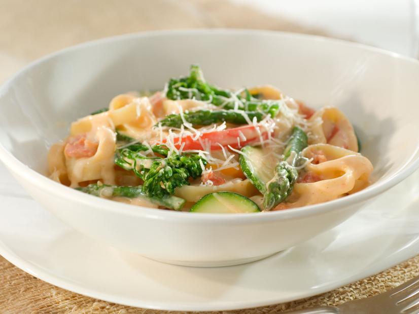 A plain white boat shaped bowl containing fettuccine topped with garden vegetables and cheese